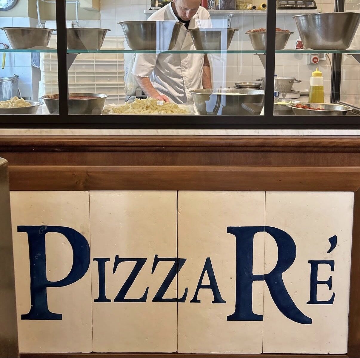 Insegna PizzaRé dipinta a mano sulle piastrelle in ceramica, con il pizzaiolo al banco sullo sfondo